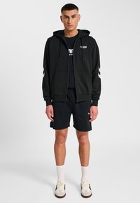 Zwarte zip-up hoodie met witte strepen op de mouwen, zwart T-shirt, zwarte shorts en crème sneakers, gedragen door een mannelijke model tegen een effen achtergrond.