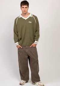 Olívazöld bordázott pulóver V-nyakkivágással és fehér részletekkel, laza barna cargo nadrággal párosítva. A megjelenést terepmintás sportcipők teszik teljessé.