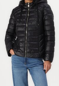 Zwarte pufferjack met capuchon, voorzien van een rits aan de voorkant, gequilt textuur en trekkoordaccenten. Draagt over blauwe denim jeans.