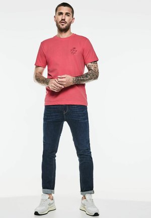 T-shirt en coton rouge à manches courtes, col rond, avec un petit motif sur la poitrine gauche, associée à un jean bleu foncé et des baskets blanches.