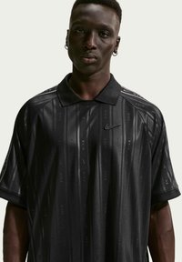 Νεαρός άνδρας που φοράει μαύρο πόλο μπλουζάκι Nike με διακριτικές κάθετες ρίγες και λογότυπα, στέκεται μπροστά σε απλό φωτεινό φόντο.