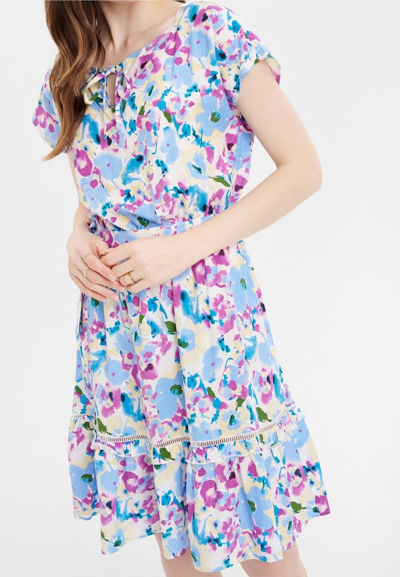 Femme portant une robe à fleurs à manches courtes avec des fleurs bleues, violettes et vertes sur un fond clair, les mains légèrement jointes à la taille.
