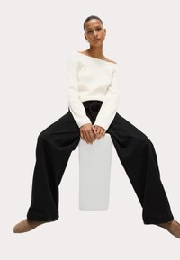 Vit, off-shoulder tröja med texturerad stickning, kombinerad med lösa, svarta vida byxor. Modell sitter på en vit kub.