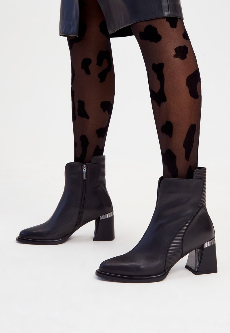 Bottines noires à talons carrés et fermetures éclair latérales portées avec des collants noirs transparents ornés de grandes taches léopard noires.
