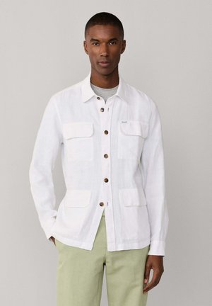 Hombre con chaqueta blanca abotonada con cuatro bolsillos delanteros sobre camisa gris y pantalones verde claro, de pie frente a un fondo liso.