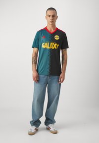 Camiseta de fútbol verde y negra con acentos rojos, que presenta "GALAXY" en texto amarillo, combinada con jeans azul claro y zapatillas blancas.