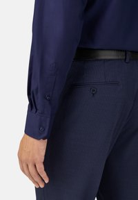 Navy-blaues Hemd mit langen Ärmeln und Knopfmanschetten, kombiniert mit dunkel gestreiften Hosen, die über eine Rückentasche und einen schwarzen Gürtel verfügen.