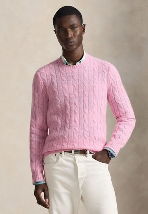 Homme portant un pull rose torsadé sur une chemise à carreaux bleue, un pantalon blanc et une montre-bracelet, debout avec une main dans la poche.