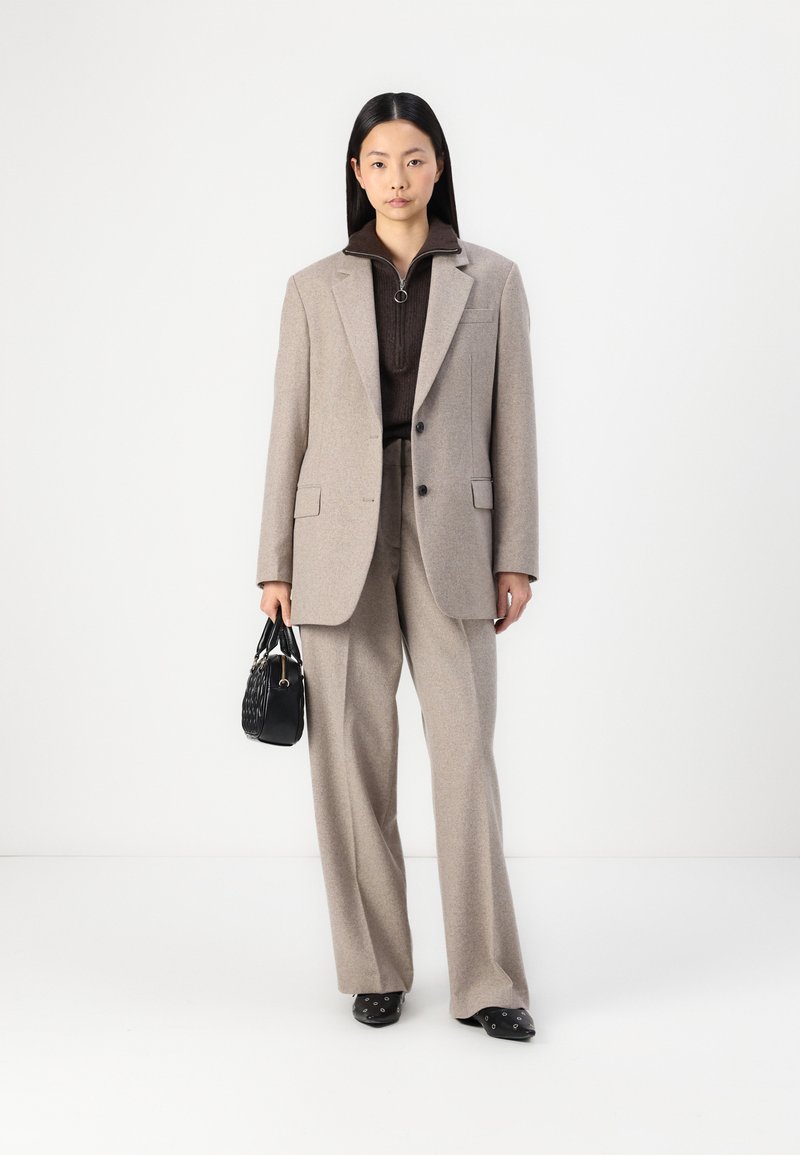 Blazer beige su misura sopra un maglione marrone a coste, abbinato a pantaloni beige a gamba larga e una borsa nera. Tacchi eleganti con dettagli in argento.