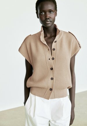 Frau mit dunkler Haut, die eine beige kurzärmelige Strickweste mit Knöpfen und weiße hoch taillierte Hose trägt, steht mit den Händen in den Taschen.
