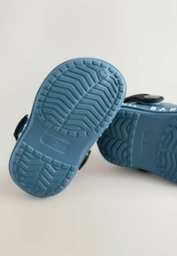 Modré gumové sandále s ryhovanou podrážkou, ktoré majú textúrovaný vzor pre lepšiu priľnavosť. Nastaviteľný čierny popruh s okrúhlou sponou.