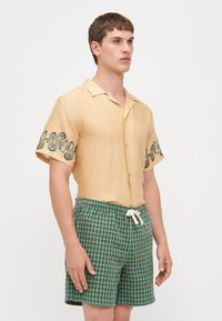 Camicia di lino gialla a maniche corte con ricamo verde a spirale sulle maniche, abbinata a pantaloni corti a quadretti verdi con cintura regolabile.