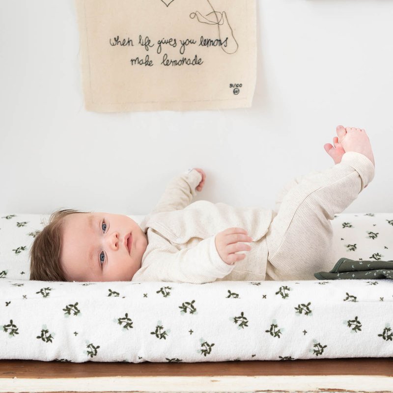 Hvid puslepude med grønne plante mønstre, beige bomuldsone-piece, baby liggende med fødderne hævet, vægdekoration med sort tekst ovenfor.