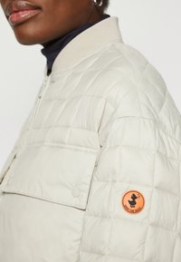 Lys beige quiltet jakke med glidelås, ribbet krage, brystlomme og en oransje rund logo-patch med silhuett av enAnd.