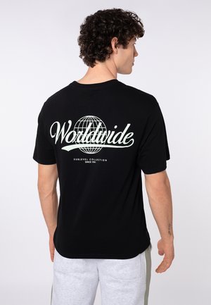 Man with curly hair wearing a black t-shirt with "Worldwide" and a globe graphic on the back, paired with light gray shorts.