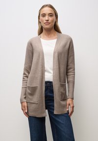 Kobieta ubrana w beżowy, otwarty kardigan z kieszeniami, na białej koszuli i niebieskich dżinsach, stojąca na tle jednolitego białego tła.