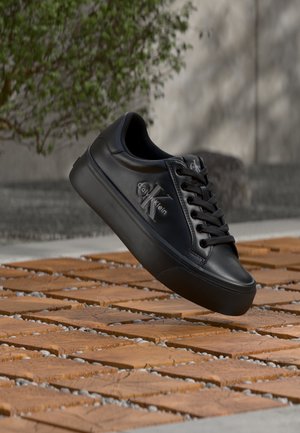 Μαύρο δερμάτινο αθλητικό παπούτσι Calvin Klein με μαύρα κορδόνια που αιωρείται πάνω από ξύλινο μπλοκ και χαλικώδη έδαφος σε εξωτερικό χώρο.