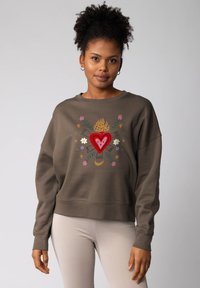 Vrouw met krullend haar in een bruine sweatshirt met een kleurrijk ontwerp van een hart, vlam, bloemen, sterren en een halve maan, gecombineerd met lichte geribde broek.
