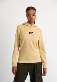 Kaptur w jasnobrązowym, teksturowanym materiale; z centralną czarną łatą z logo. W połączeniu z ciemnymi spodniami, prezentującymi luźny krój.