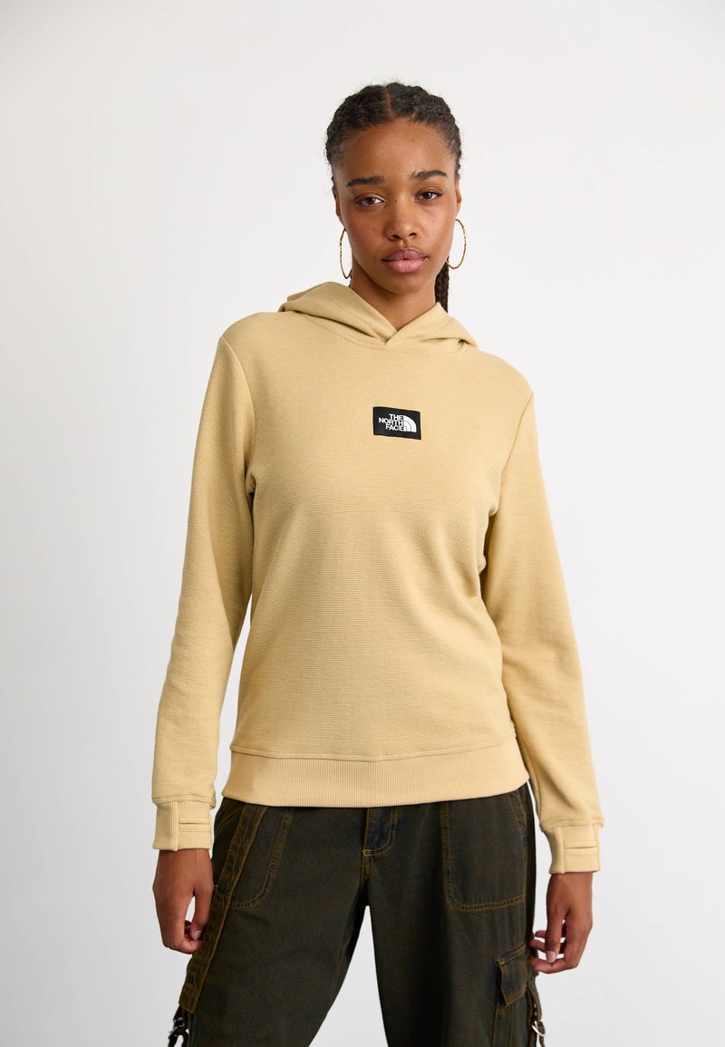 Kaptur w jasnobrązowym, teksturowanym materiale; z centralną czarną łatą z logo. W połączeniu z ciemnymi spodniami, prezentującymi luźny krój.