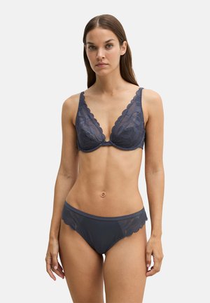 Woman with long brown hair wearing dark gray lace bra and matching lace-trimmed underwear standing against plain light background.