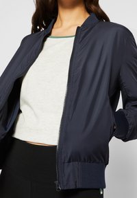 Blouson aviateur bleu marine avec fermeture éclair frontale, poignets côtelés et texture lisse et brillante. Associé à un haut gris clair et un pantalon noir.