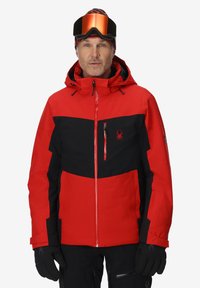 Veste de ski rouge et noire avec fermeture éclair sur le devant, capuche ajustable et plusieurs poches zippées. Présente un logo sur la poitrine et un tissu texturé.