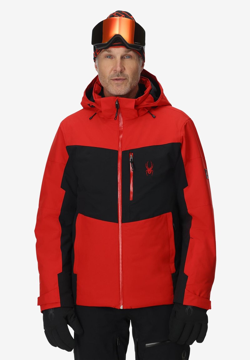 Veste de ski rouge et noire avec fermeture éclair sur le devant, capuche ajustable et plusieurs poches zippées. Présente un logo sur la poitrine et un tissu texturé.