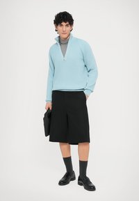 Pull-over bleu clair côtelé avec demi-fermeture éclair, col roulé gris en dessous, short noir, chaussettes foncées et chaussures noires. Tenant une veste noire.