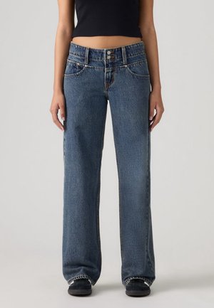 Personne portant un jean denim bleu ample avec une taille à double bouton et des baskets noires, debout sur un fond clair uni.