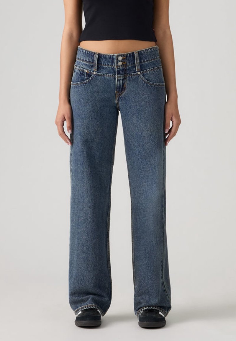 Personne portant un jean denim bleu ample avec une taille à double bouton et des baskets noires, debout sur un fond clair uni.
