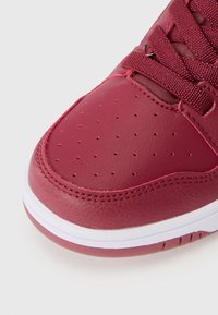 Punane nahast sneaker, millel on aukudega varvaste osa, tekstuuriga paelad ja valge kummine tald, millel on peen roosa aktsent.