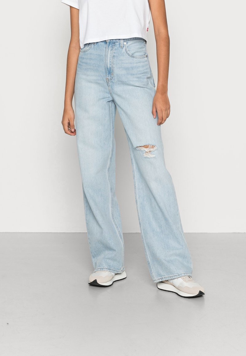 Personne portant un jean taille haute large couleur bleu clair avec une déchirure sur la cuisse droite, un crop top blanc et des baskets beige, debout sur un sol gris.