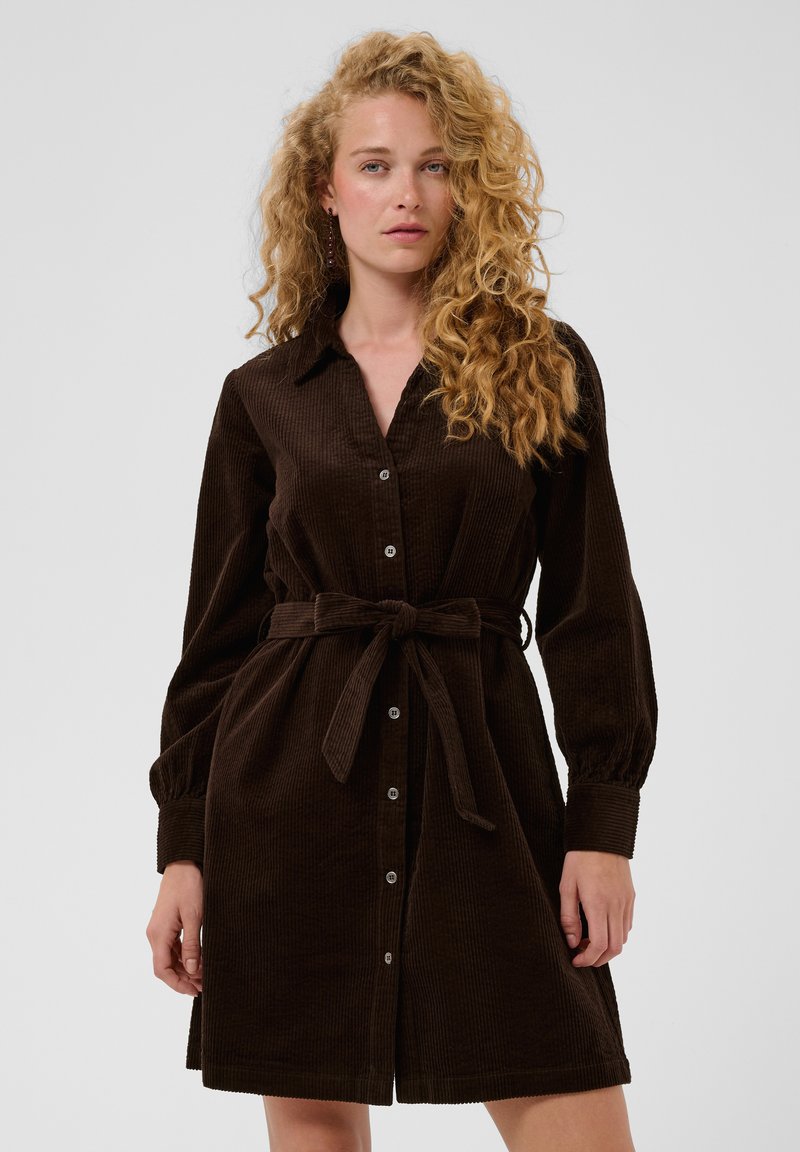 Femme aux longs cheveux blonds bouclés portant une robe en velours côtelé marron foncé à manches longues avec des boutons et une ceinture nouée à la taille.