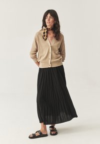 Cardigan lavorato a maglia beige con scollo a V e chiusura a bottoni, abbinato a una gonna maxi plissettata nera. Sandali neri e una sciarpa a pois completano l'outfit.