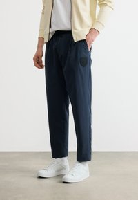 Άνδρας που φοράει μπλε παντελόνια με μπαλώματα, λευκά αθλητικά παπούτσια, λευκές κάλτσες, άσπρο πουκάμισο και μπεζ μπουφάν με φερμουάρ, στέκεται σε τσιμεντένιο πάτωμα.