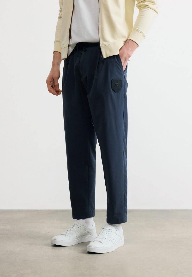 Άνδρας που φοράει μπλε παντελόνια με μπαλώματα, λευκά αθλητικά παπούτσια, λευκές κάλτσες, άσπρο πουκάμισο και μπεζ μπουφάν με φερμουάρ, στέκεται σε τσιμεντένιο πάτωμα.