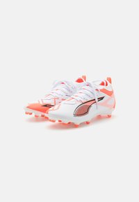 Botas de fútbol blancas con acentos en naranja, empeine sintético texturizado, logo negro y múltiples tapones moldeados en la suela para tracción.