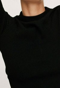 Gros plan sur une personne portant un pull en maille côtes noir à col rond haut, bras levés partiellement visibles.