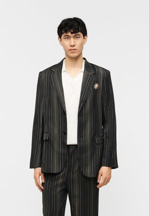 Jeune personne portant un costume noir à rayures fines dorées, une chemise blanche et une broche en forme de soleil sur le revers de la veste, debout devant un fond blanc.