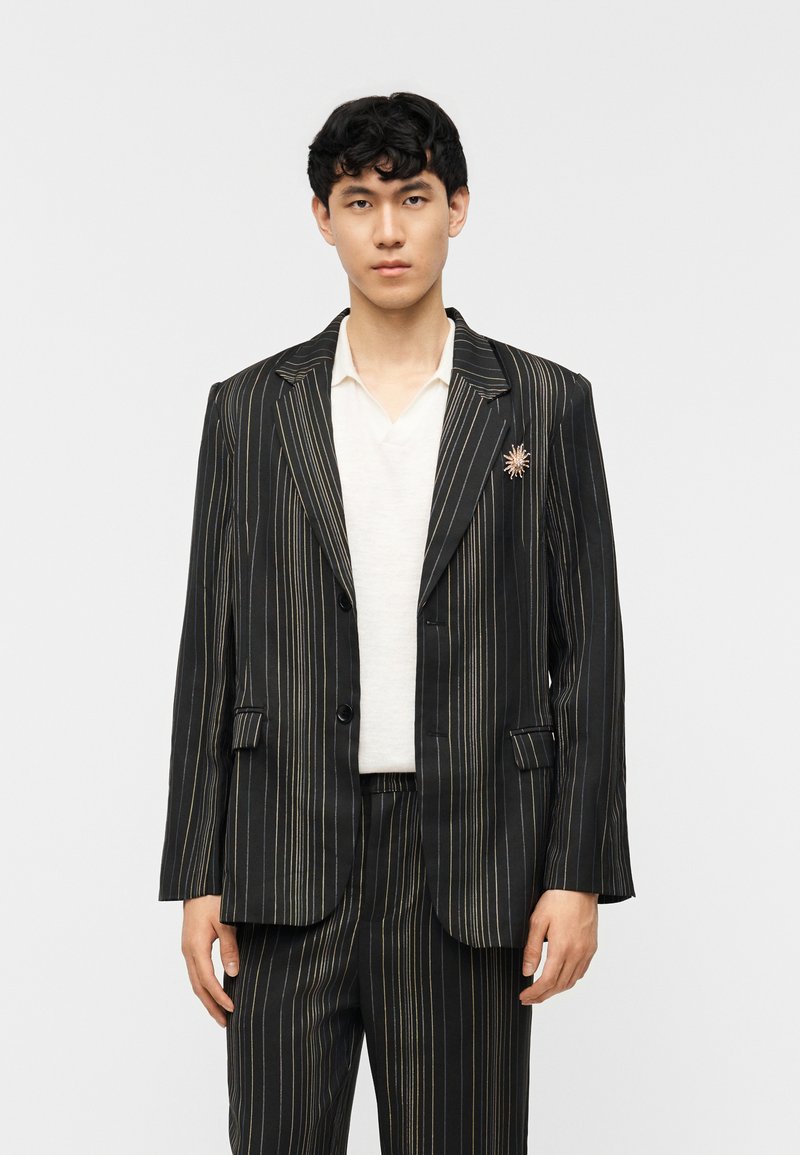 Νεαρό άτομο φοράει μαύρο κοστούμι με λεπτές χρυσές ρίγες, λευκό πουκάμισο και καρφίτσα σε σχήμα ήλιου στο πέτο του σακακιού, στέκεται μπροστά σε λευκό φόντο.