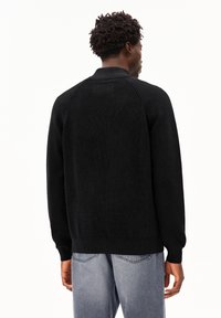 Man in een zwart gebreid sweater met lange mouwen en lichtblauwe jeans, met zijn rug naar ons gekeerd, waarbij de textuur van de stof en de geribbelde halslijn zichtbaar zijn.