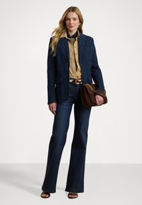 Denim blazer en flared jeans in donkerblauw. Lichtbruine overhemd met knopen eronder, bruine handtas met kettinghengsel, en bruine sandalen.