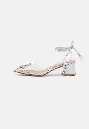 Sandale à talon bloc argenté avec bride transparente sur les orteils, ornement en strass et lacets à nouer autour de la cheville sur fond blanc.