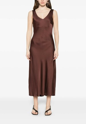 Donna che indossa un abito midi marrone in raso senza maniche con scollo a V e sandali neri open-toe, in piedi su sfondo bianco.