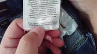 Etiqueta em roupa de denim mostrando instruções de cuidado, composição do tecido e país de fabricação. Mão segurando a peça perto da cintura.