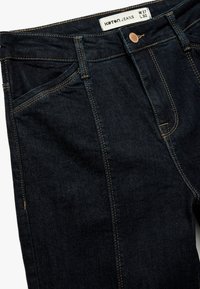 Donker gewassen denim jeans met contrasterende gouden stiksels, vijf-pocket ontwerp en een koperen knoop. Label leest "KOTAN JEANS W 27 L 32."