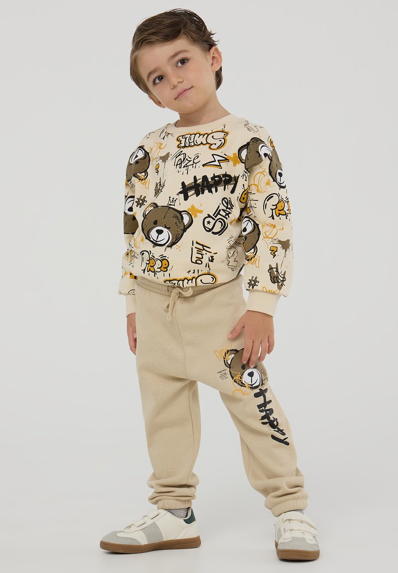 Beżowy dres dla dzieci z wzorzystą kremową bluzą z grafiką niedźwiedzia oraz nadrukiem, w komplecie z dopasowanymi beżowymi spodniami.