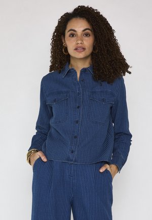 Donna con capelli ricci che indossa una camicia a righe blu con bottoni e pantaloni abbinati, mani in tasca, accessoriata con orecchini e braccialetti d'oro.