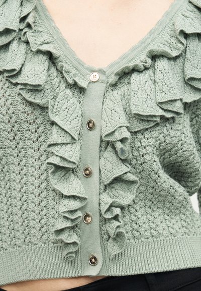 Lichtgroene gebreide cardigan met geruite randen en vijf knopen aan de voorkant, gedragen over blote huid.